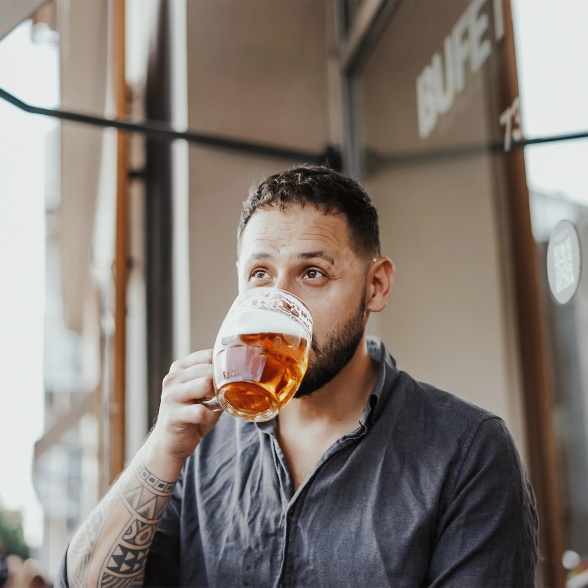Peter Zošiak sedí na venkovní terase restaurace a pije pivo z velkého skleněného půllitru. Má tmavě modrou košili, tetování na ruce a zasněný pohled. V pozadí je nápis „BUFET“.