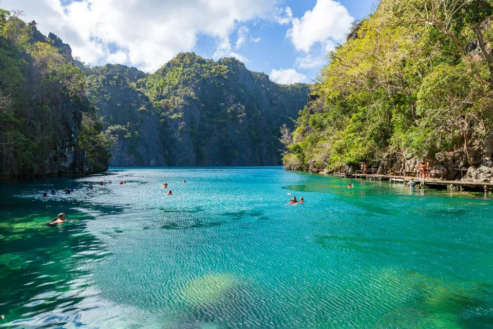 Turisté plavající v křišťálově čisté tyrkysové vodě jezera Kayangan poblíž ostrova Coron na Filipínách. Jezero je obklopeno dramatickými vápencovými útesy pokrytými svěží vegetací, což vytváří odlehlé a malebné přírodní prostředí. Dřevěná promenáda na pravé straně poskytuje přístup k vodě, kde si návštěvníci užívají relaxaci a objevování krás okolní krajiny. Sluneční světlo pronikající skrz stromy zvýrazňuje syté modré a zelené odstíny vody.