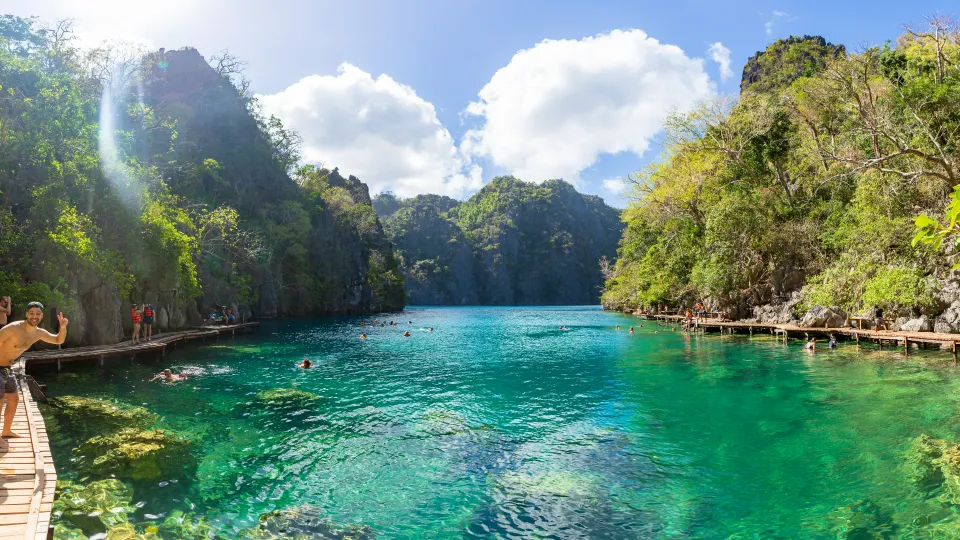 Turisté plavající v křišťálově čisté tyrkysové vodě jezera Kayangan poblíž ostrova Coron na Filipínách. Jezero je obklopeno dramatickými vápencovými útesy pokrytými svěží vegetací, což vytváří odlehlé a malebné přírodní prostředí. Dřevěná promenáda na pravé straně poskytuje přístup k vodě, kde si návštěvníci užívají relaxaci a objevování krás okolní krajiny. Sluneční světlo pronikající skrz stromy zvýrazňuje syté modré a zelené odstíny vody.