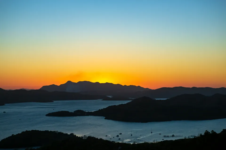 Panoramatický pohled na ostrov Coron na Filipínách při západu slunce. Slunce zapadá za horský horizont, vytvářející nádherný přechod barev od sytě oranžové po jemně modrou oblohu. Pod siluetou hor se rozprostírá moře s malými ostrovy a kotvícími loděmi. Klidná voda odráží poslední zbytky denního světla, čímž dodává scenérii poklidnou a magickou atmosféru tropického ráje.