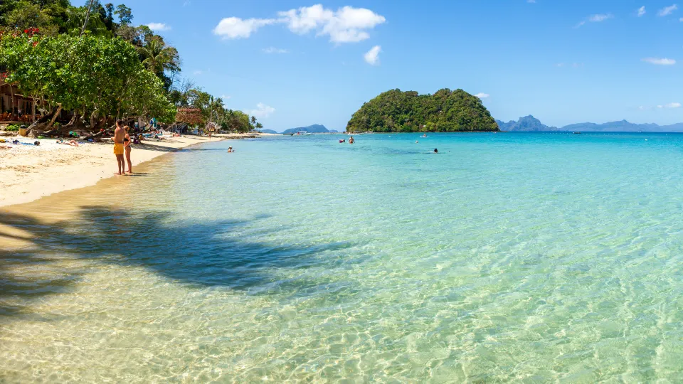 Pohled na křišťálově čisté vody Las Cabañas Beach na Filipínách s jemným písčitým pobřežím, kde se lidé procházejí a relaxují. V pozadí se rýsuje malý zelený ostrov obklopený tyrkysovým mořem. Slunečné počasí a modrá obloha dodávají snímku letní tropickou atmosféru.