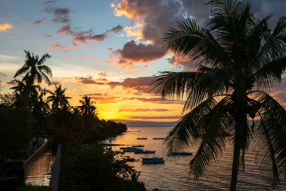Západ slunce nad mořem u hotelu Nine Degrees North Pavilon na ostrově Panglao, Filipíny. Siluety palem a lodí na vodní hladině vytvářejí tropickou atmosféru, zatímco obloha hraje odstíny oranžové, růžové a modré.