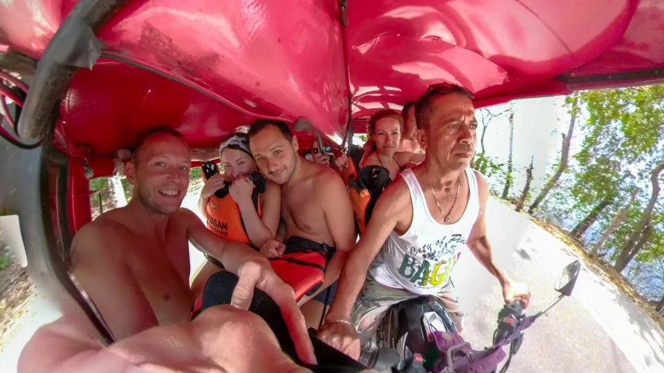 Skupina turistů namačkaná v malém červeném motorizovaném tuk-tuku během cesty na canyoning v oblasti Kawasan Falls na Filipínách. Řidič v bílém tílku soustředěně řídí, zatímco pasažéři, někteří v záchranných vestách, se smějí a pořizují selfie. Stísněný prostor a tropické vedro vytvářejí autentickou atmosféru cestování po Filipínách.
