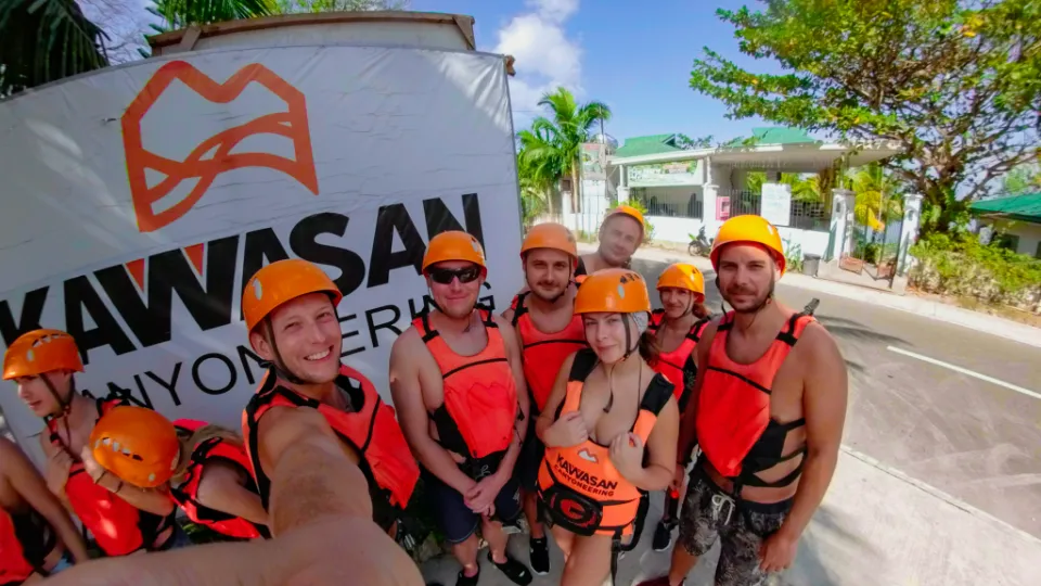 Skupina dobrodruhů ve výrazných oranžových helmách a záchranných vestách pózuje pro selfie před bannerem „Kawasan Canyoneering“ na Filipínách. Všichni se usmívají a připravují se na adrenalinový zážitek v tropické přírodě. V pozadí jsou palmy, modrá obloha a budovy s zelenými střechami, vytvářející typickou atmosféru filipínské vesnice.