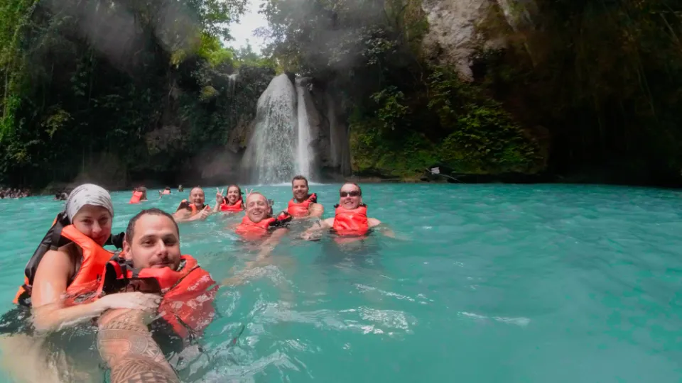 Skupina turistů plave v tyrkysově modré vodě pod vodopádem Kawasan Falls na Filipínách. Všichni mají oranžové záchranné vesty a užívají si osvěžující koupel v tropickém ráji. Hustá džungle a skalní stěny obklopují vodopád, jehož proud se řítí do laguny. Úsměvy a radost ve tvářích zachycují nezapomenutelný okamžik dobrodružství.