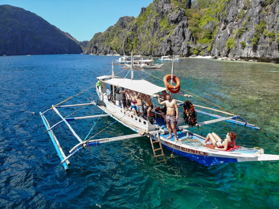 Tradiční filipínská outrigger loď (banka) s turisty kotví u Matinloc Shrine poblíž El Nido na ostrově Palawan, Filipíny. Jeden muž stojí na boku lodi a drží záchranný kruh, zatímco ostatní relaxují, povídají si nebo se připravují ke koupání. Žena leží na přídi a užívá si sluneční paprsky. V pozadí jsou dramatické vápencové útesy a křišťálově čistá voda, která odhaluje korálové útesy pod hladinou. Tropická scenérie zachycuje krásu filipínského ostrovního dobrodružství.