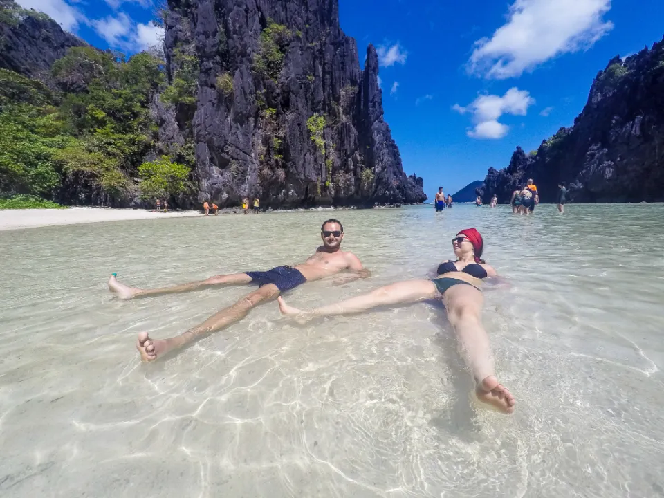 Dva turisté relaxují v mělké, křišťálově čisté vodě Hidden Beach v El Nido na ostrově Palawan, Filipíny. Obklopeni strmými vápencovými útesy si užívají tropické slunce pod jasně modrou oblohou s několika bílými mraky. V pozadí jsou další návštěvníci, kteří se procházejí po písku a laguně. Scenérie zachycuje nádhernou a odlehlou atmosféru této skryté pláže.