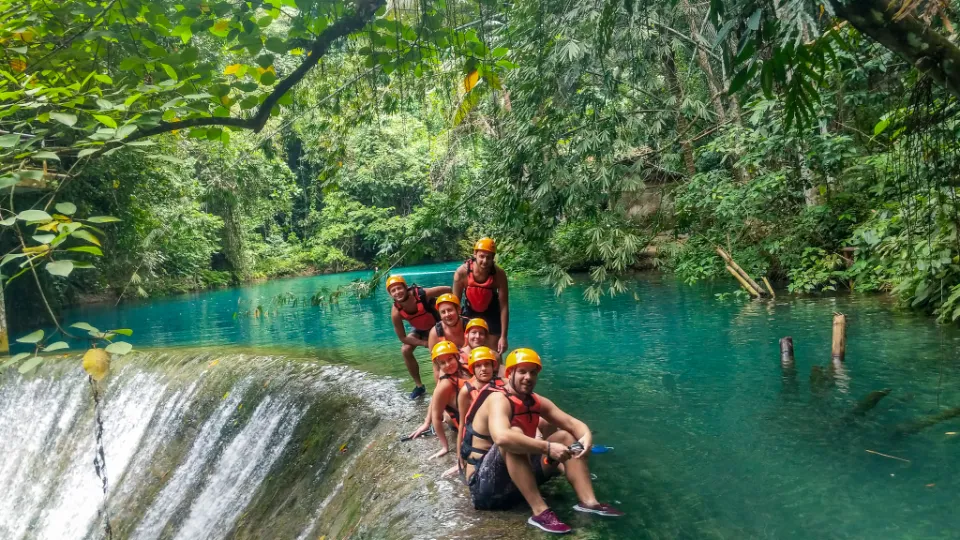 Skupina dobrodruhů v oranžových helmách a záchranných vestách sedí na okraji malého vodopádu během canyoningu v oblasti Kawasan Falls na Filipínách. Tyrkysově modrá řeka obklopená hustou tropickou džunglí vytváří nádhernou přírodní scenérii. Nad skupinou se sklání větve stromů, zatímco voda klidně proudí přes skalní hranu. Atmosféra je dobrodružná a zároveň klidná, zachycující krásu a vzrušení z objevování této oblíbené filipínské destinace.