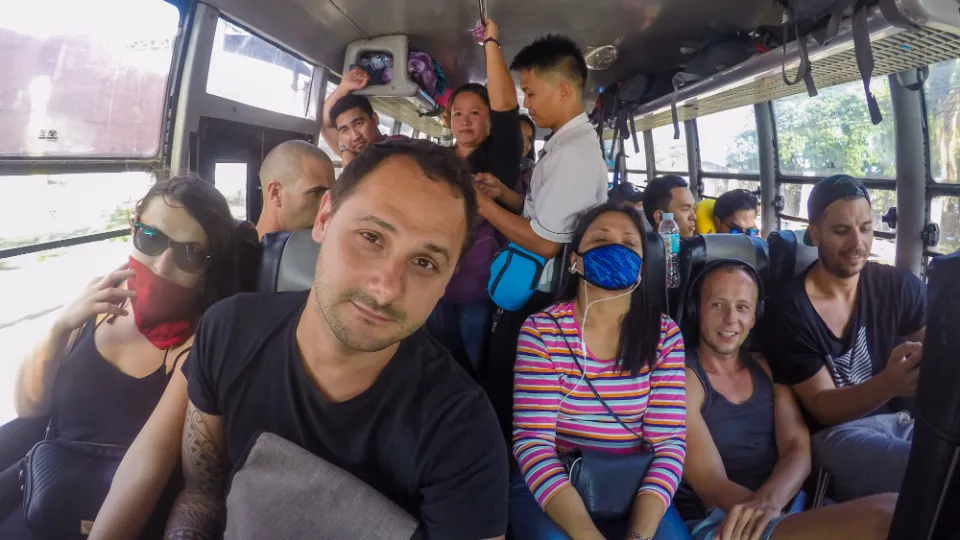 Přeplněný autobus na Filipínách s cestujícími sedícími i stojícími v úzkém prostoru. Muž v popředí pořizuje selfie, zatímco ostatní pasažéři kolem něj odpočívají, poslouchají hudbu nebo sledují své telefony. Žena v barevném pruhovaném tričku s maskou na obličeji spí opřená o sedadlo. Někteří cestující drží zavazadla nebo se přidržují madel. Sluneční světlo proniká okny, odhalující pohybující se krajinu venku. Atmosféra zachycuje typickou scénu veřejné dopravy na Filipínách.