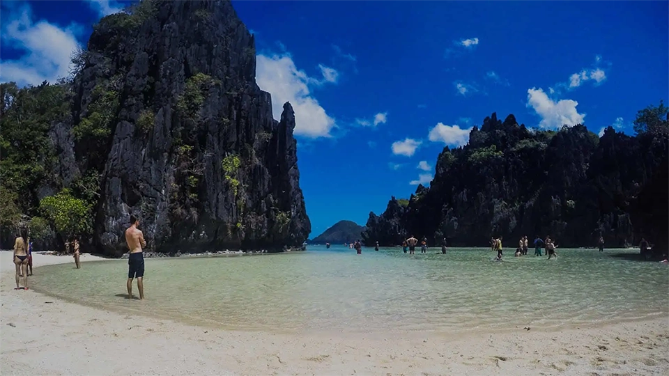 Hidden Beach v El Nido na ostrově Palawan, Filipíny. Pláž s jemným bílým pískem je obklopena impozantními vápencovými útesy, které se tyčí nad tyrkysově modrou vodou. Turisté se procházejí mělkou lagunou směrem k otevřenému moři, zatímco v dálce je vidět další ostrov. Slunečné počasí a jasně modrá obloha s několika bílými mraky dodávají scenérii tropický a exotický vzhled.