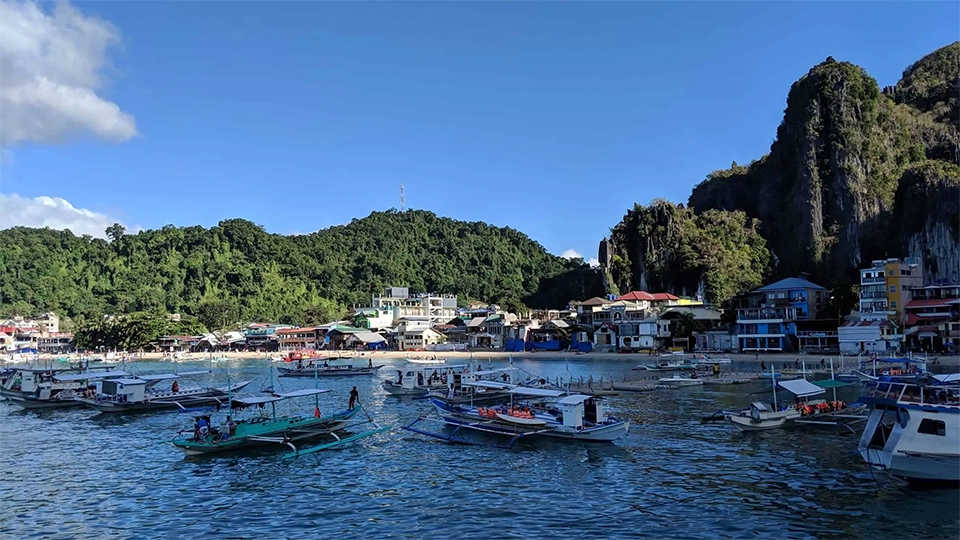 Přístavní město El Nido na ostrově Palawan na Filipínách s tradičními filipínskými outrigger loděmi (bangkami) zakotvenými ve vodě. Na pozadí se tyčí dramatické vápencové útesy a zelené kopce, zatímco pobřeží lemují barevné budovy, hotely a restaurace. Slunečné počasí a modrá obloha dodávají tropickou atmosféru tomuto oblíbenému výchozímu bodu pro ostrovní výlety po souostroví Bacuit.