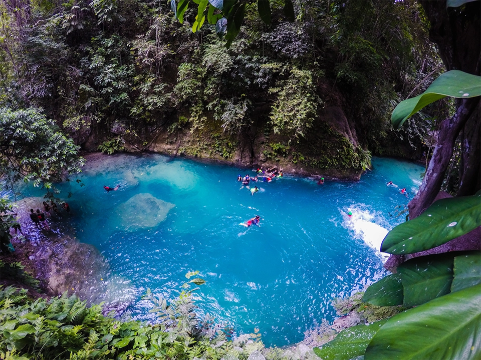 Turisté v záchranných vestách plavou a skáčou do tyrkysově modré vody během canyoningu v oblasti Kawasan Falls na Filipínách. Vodopád na pravé straně vytváří bílou pěnu, zatímco kolem něj se rozprostírá hustá tropická vegetace. Skály pokryté mechem a visící rostliny dodávají scenérii divoký a exotický vzhled. Lidé na břehu i ve vodě si užívají adrenalinový zážitek v této přírodní oáze, která je známá svou průzračnou vodou a malebnou džunglí.