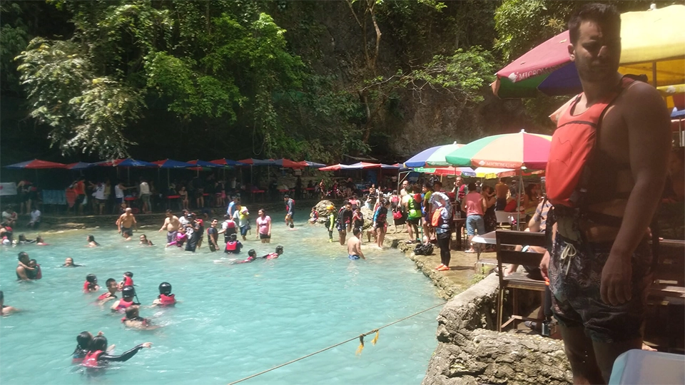 Rušná oblast u Kawasan Falls na Filipínách plná turistů a místních obyvatel. Lidé v záchranných vestách si užívají koupání v tyrkysově modré vodě, zatímco ostatní relaxují pod barevnými slunečníky a navštěvují stánky s jídlem a suvenýry. Tropická atmosféra a živá energie místa odrážejí popularitu této přírodní atrakce.