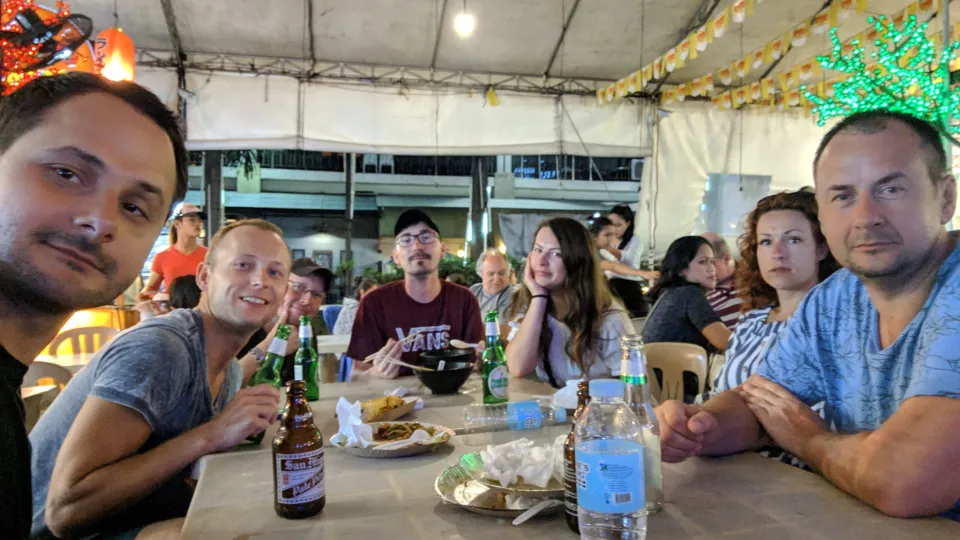 Skupina přátel sedí u stolu v nočním podniku v Makati, Manila, Filipíny. Na stole jsou lahve piva San Miguel, voda a talíře s jídlem. V pozadí jsou další hosté, světelné dekorace a stánky s jídlem, které vytvářejí živou atmosféru.