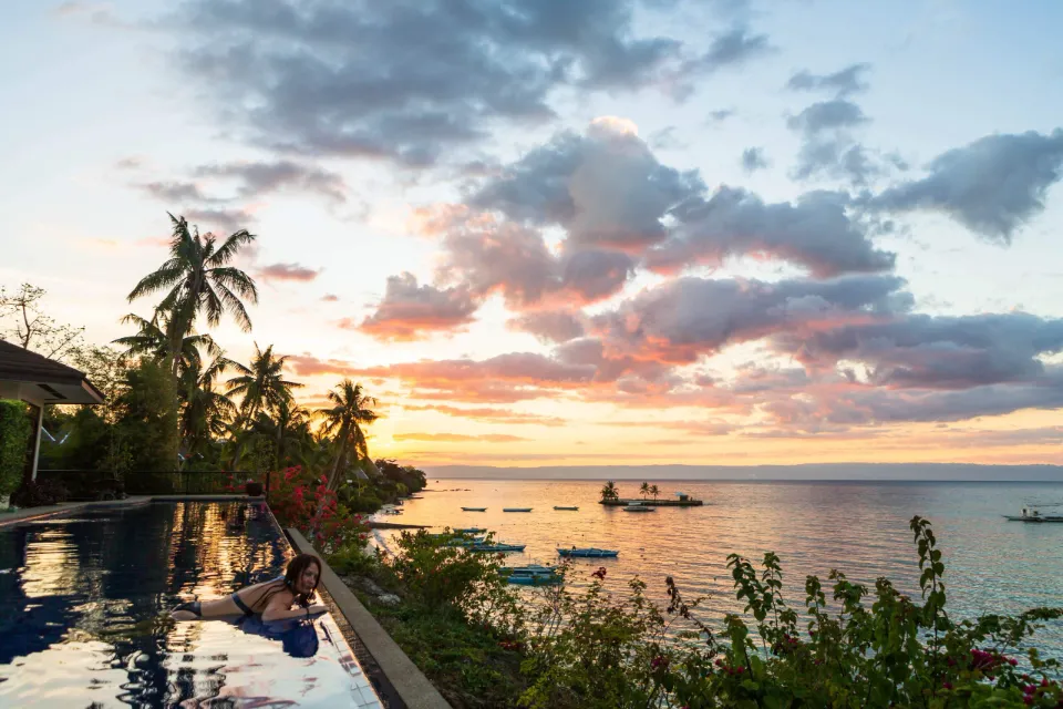 Malebný západ slunce nad mořem v hotelu Nine Degrees North Pavilion na ostrově Panglao. Žena leží na okraji infinity bazénu, zatímco v pozadí jsou tropické palmy, rozkvetlé keře a klidná vodní hladina s kotvícími loďkami.