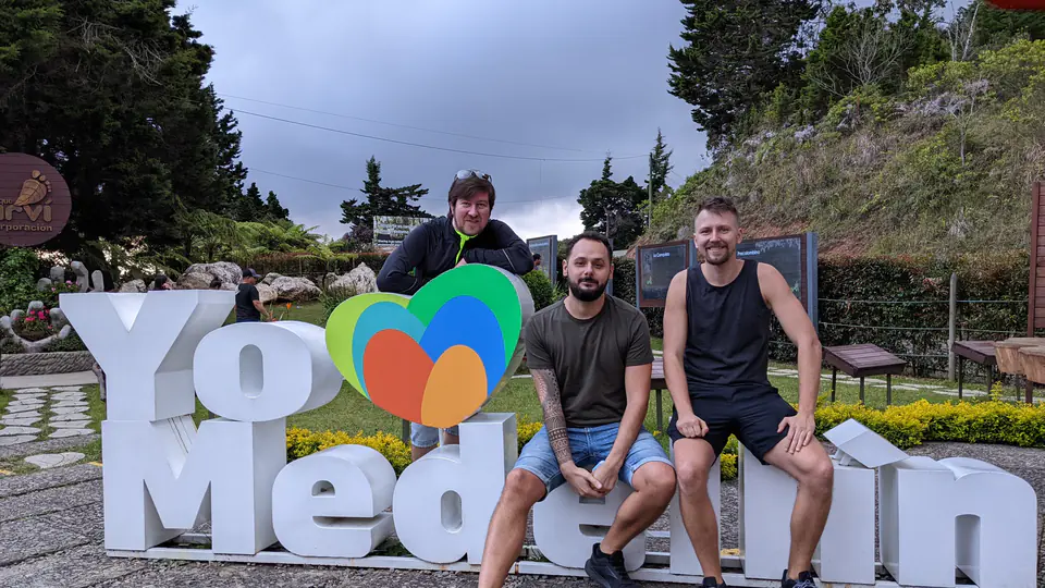 Tři muži pózují u barevného nápisu "Yo ❤️ Medellín" v přírodním parku poblíž Medellínu v Kolumbii. Jeden muž se opírá o písmeno "Y", zatímco další dva sedí před nápisem. V pozadí je vidět zelená vegetace, informační tabule a oblačné nebe. Atmosféra působí příjemně a výletně.