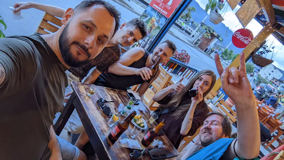 Skupina šesti lidí sedí u dřevěného stolu v baru nebo restauraci v Guatapé, Kolumbie. Pijí pivo a tequilu, smějí se, dělají gesta do kamery a užívají si přátelský večer. Stůl je pokrytý lahvemi, skleničkami a talířkem s limetkami. V pozadí je vidět ulice a město pod horami. Celá scéna působí uvolněně a radostně.
