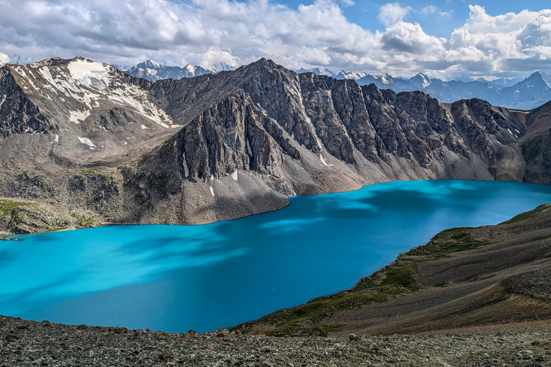 Rozsáhlý výhled na tyrkysové jezero Ala-Kul v Kyrgyzstánu s impozantním horským pásem v pozadí. Drsné skalnaté vrcholy s ledovými čepicemi dodávají krajině epickou atmosféru. Obloha je kombinací modré a zatažených oblastí, přičemž světlo vytváří nádherné odrazy na hladině jezera.