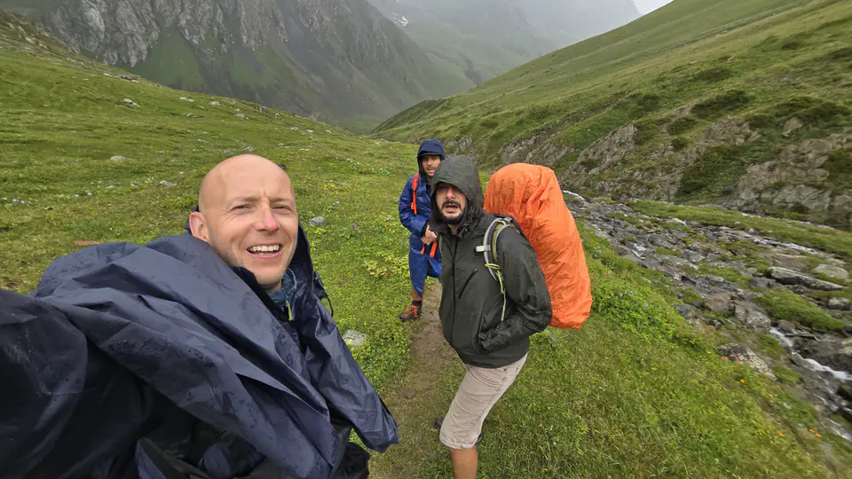 Tři turisté na horské stezce během deštivého dne, obklopení zelenými loukami a skalnatými svahy. Muž v popředí má na sobě tmavý neprůstřelný plášť a usmívá se do fotoaparátu. Druhý turista, v zelené bundě s kapucí, nese batoh chráněný oranžovým pláštěm. Třetí turista, oblečený v modrém nepromokavém plášti, stojí dále v pozadí. Vedle nich teče horský potok a atmosféra je mlhavá, což naznačuje sychravé počasí.