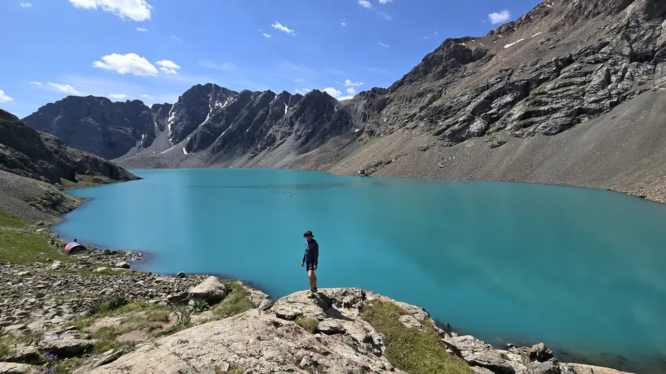 Malebné tyrkysové jezero Ala-Kul obklopené strmými horskými hřbety v Kyrgyzstánu. V popředí stojí turista na skále, obdivující nádhernou přírodu. Jezero má křišťálově čistou vodu, která odráží modrou oblohu. Na břehu jsou viditelné stany, což naznačuje kempování v této odlehlé oblasti.