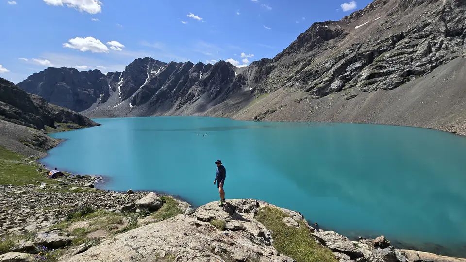 Malebné tyrkysové jezero Ala-Kul obklopené strmými horskými hřbety v Kyrgyzstánu. V popředí stojí turista na skále, obdivující nádhernou přírodu. Jezero má křišťálově čistou vodu, která odráží modrou oblohu. Na břehu jsou viditelné stany, což naznačuje kempování v této odlehlé oblasti.