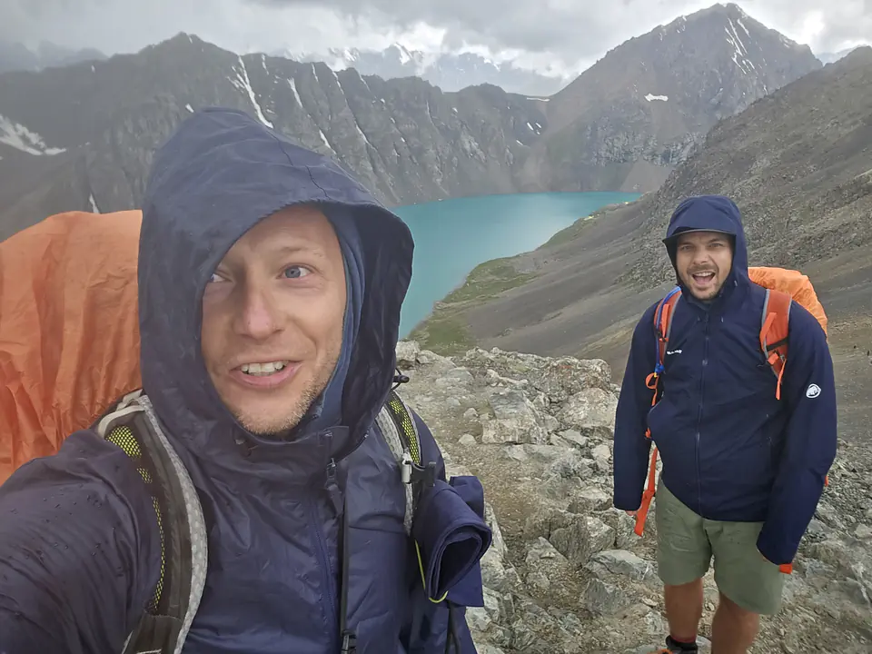 Dva turisté v nepromokavých bundách stojí na skalnatém hřebeni s výhledem na tyrkysové jezero Ala-Kul v Kyrgyzstánu. Obloha je zatažená tmavými mraky, což naznačuje blížící se bouři. Jeden turista se usmívá, zatímco druhý dokumentuje moment. Strmé hory v pozadí dodávají dramatický nádech scenérii.