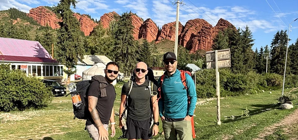 Tři turisté pózují na zelené louce před červenými skalními útvary v oblasti Jeti-Ögüz v Kyrgyzstánu. V pozadí je vidět dřevěné domy, stromy a modrá obloha s rozházenými mraky. Muži mají turistické oblečení a batohy, připraveni na dobrodružství v přírodě.