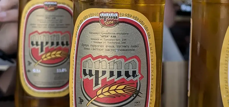 Detailní záběr tří lahví piva Arpa z Kyrgyzstánu na lesklém stole. Etikety na lahvích mají zlatý motiv ječmene s cyrilicí, uvádějící obsah alkoholu 11,0 %. Na etiketách je výrazně zobrazen název značky „Arpa“ a zmínka o původu piva. Na jedné z lahví je připevněná daňová nálepka na hrdle.