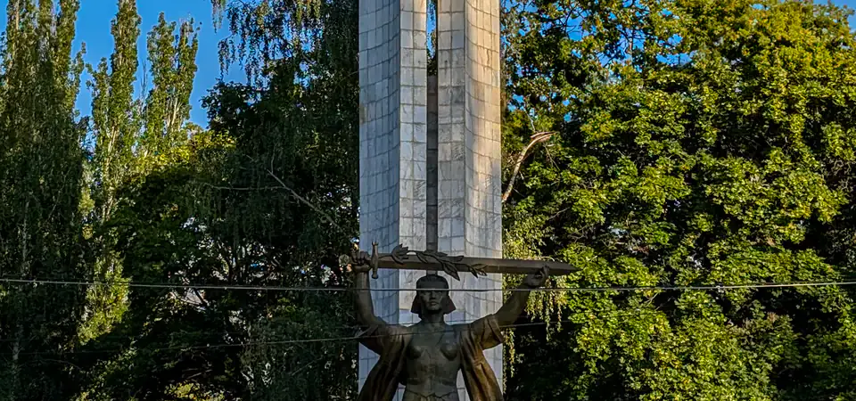 Památník z období Sovětského svazu se svislou mramorovou konstrukcí a bronzovým emblémem kosáka a kladiva u vrcholu. Pod tímto symbolem stojí bronzová socha ženy, která drží meč zvednutý oběma rukama. Památník je obklopen stromy a dlažbou, kde sedí několik lidí na schodech.