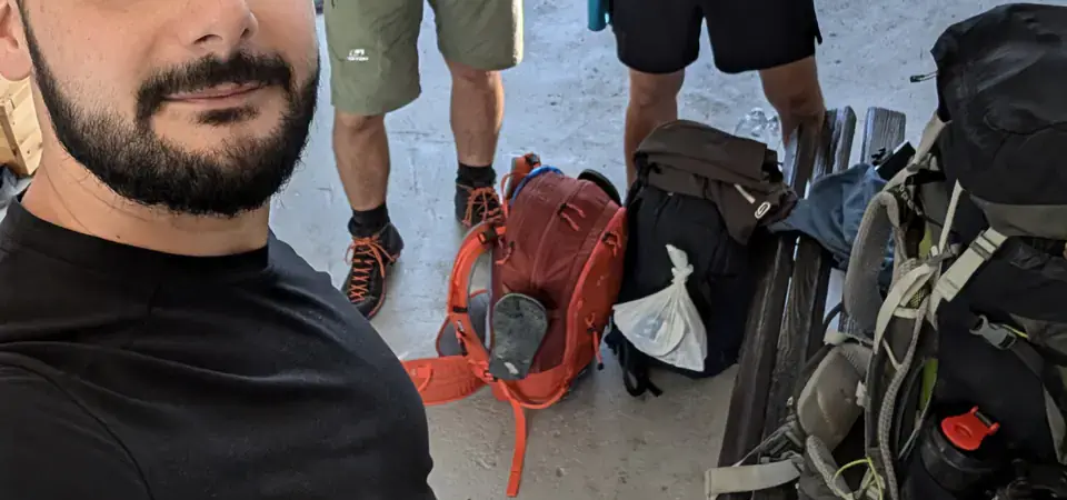 Tři muži uvnitř přístřešku se skleněnými stěnami a dřevěnou konstrukcí. Jeden z nich dělá selfie, zatímco další dva pózují v pozadí s úsměvy. Na lavici a podlaze jsou rozložené turistické batohy a vybavení. Světlo proniká dovnitř, naznačující brzké ranní hodiny.