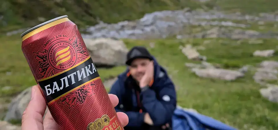 Osoba drží plechovku piva Baltika 9 s červeným designem a zlatými detaily v ruce v popředí. Na plechovce je viditelný ruský nápis „БАЛТИКА 9 КРЕПКОЕ СВETLOЕ ПИВО“. V pozadí se nachází turista sedící na trávě, oblečený v tmavé bundě a čepici, s opřenou hlavou o ruku. Kolem něj jsou rozložené turistické věci, včetně batohu a spacáku. Scéna se odehrává v horském údolí s trávnatými svahy, kamennými útvary a potokem, přičemž v dálce jsou viditelné vrcholy hor. Nad krajinou je modrá obloha s několika bílými mraky.