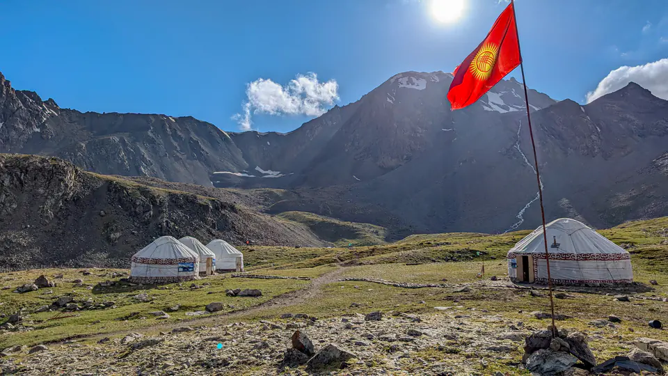 Rozsáhlý výhled na tyrkysové jezero Ala-Kul v Kyrgyzstánu s impozantním horským pásem v pozadí. Drsné skalnaté vrcholy s ledovými čepicemi dodávají krajině epickou atmosféru. Obloha je kombinací modré a zatažených oblastí, přičemž světlo vytváří nádherné odrazy na hladině jezera.