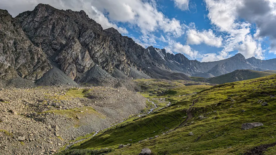 Rozsáhlé horské údolí v Kyrgyzstánu s klikatícím se potokem a dramatickými skalnatými hřbety. Sluneční paprsky osvětlují zelené svahy, zatímco nad vrcholy se vznášejí oblaky. Kamenitý terén a prudké srázy naznačují divokou a nespoutanou přírodu Střední Asie.