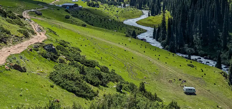Pohled na nádherné údolí Altyn-Arashan v Kyrgyzstánu s klikatícím se řekou mezi zelenými kopci a hustými jehličnatými lesy. V dálce jsou viditelné roztroušené budovy a jurty, typické pro místní pastevecké osady. Horský hřeben v pozadí dodává krajině dramatickost, zatímco modrá obloha s bílými mraky zdůrazňuje letní atmosféru této odlehlé a nedotčené přírodní oblasti.