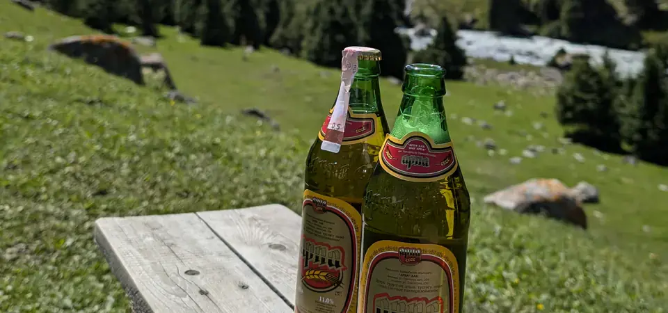 Dvě skleněné lahve kyrgyzského piva značky „Арпа“ (Arpa) položené na dřevěné lavici v přírodním prostředí. V pozadí se rozprostírá malebné horské údolí s řekou, zelenými loukami a roztroušenými jurtami mezi jehličnatými lesy. Scenérie evokuje atmosféru odpočinku a vychutnávání místních produktů v krásném horském prostředí Kyrgyzstánu.