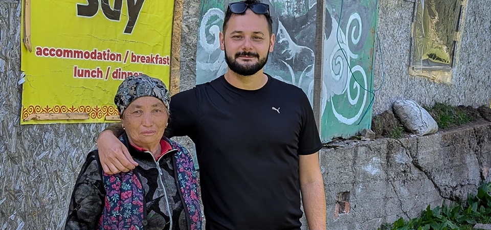 Muž v černém tričku s rukou kolem starší ženy v tradičním oděvu pózuje před budovou v kempu &quot;Yurt Camp Bulak Say&quot; v Kyrgyzstánu. Na stěně je žlutý banner s nápisem kempu a vedle něj nástěnná malba muže v tradičním oděvu. V pozadí jsou viditelné hory a jehličnaté lesy.