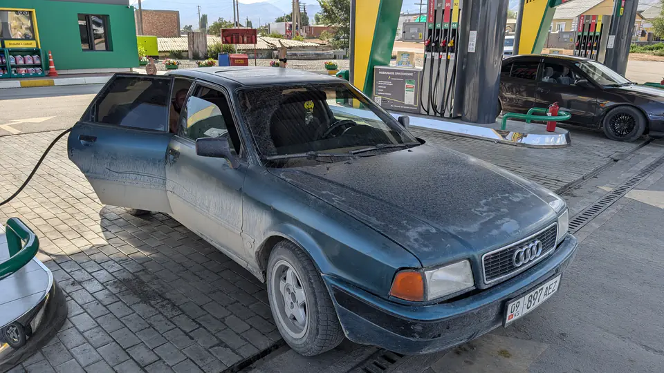 Staré, zaprášené Audi s kyrgyzskou ŠPZ &quot;KG 897 AEZ&quot; na čerpací stanici v Kyrgyzstánu. Řidič dává palec nahoru z okna, zatímco se auto tankuje. V pozadí jsou další vozidla a místní infrastruktura.