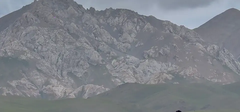 Osamělá postava zahalená v černém plášti sedí na betonové konstrukci v otevřené stepi v Kyrgyzstánu. Za ní se tyčí dramatické skalnaté hory, zatímco nad krajinou se shromažďují tmavé bouřkové mraky.