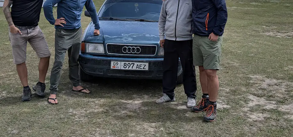 Čtyři muži pózují u starého Audi s kyrgyzskou ŠPZ na trávnaté ploše u jezera v Kyrgyzstánu. V pozadí jsou rozsáhlé hory a modrá obloha s mraky. Tato scéna zachycuje dobrodružnou atmosféru cestování po zemi.