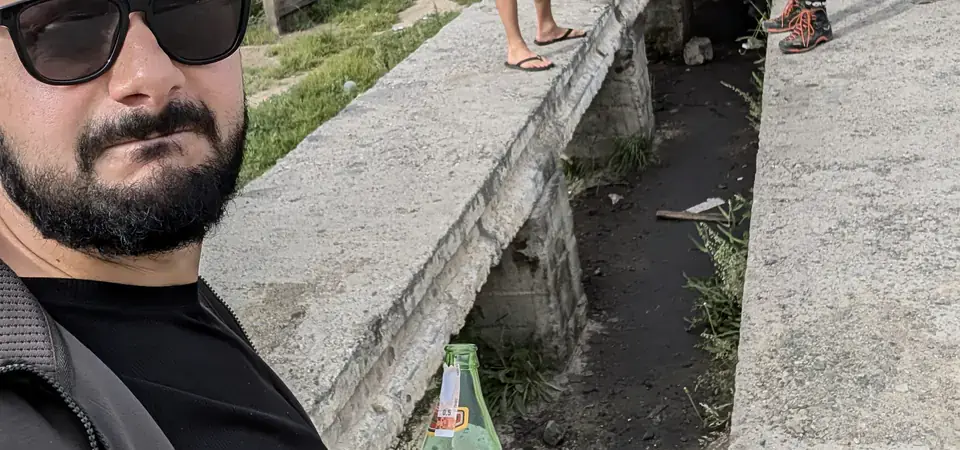 Muž v slunečních brýlích drží láhev piva, zatímco dva další stojí u zaparkovaného Audi na mostě nad příkopem ve vesnici v Kyrgyzstánu. V pozadí jsou charakteristické kopcovité hory a jednoduché venkovské domy.