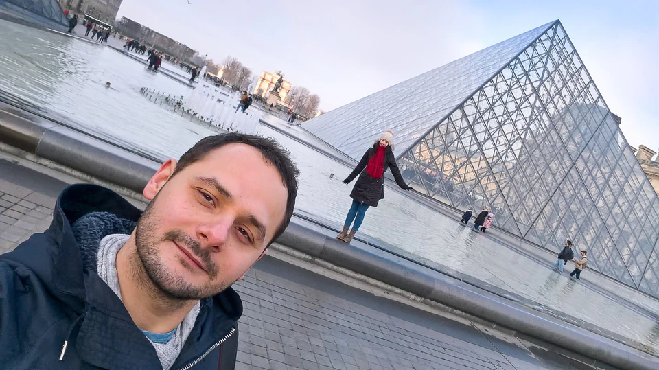 Peter Zošiak pořizuje selfie před ikonickou skleněnou pyramidou v Louvru v Paříži. Za ním je žena v červené šále pózující na okraji fontány, kolem se pohybují turisté.