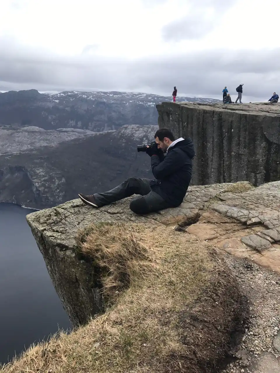 Peter Zošiak sedí na hraně dramatického skalního útesu v Norsku, fotí krajinu pod sebou. V pozadí jsou další turisté, hory a fjord. Nebe je zatažené, atmosféra je sychravá.