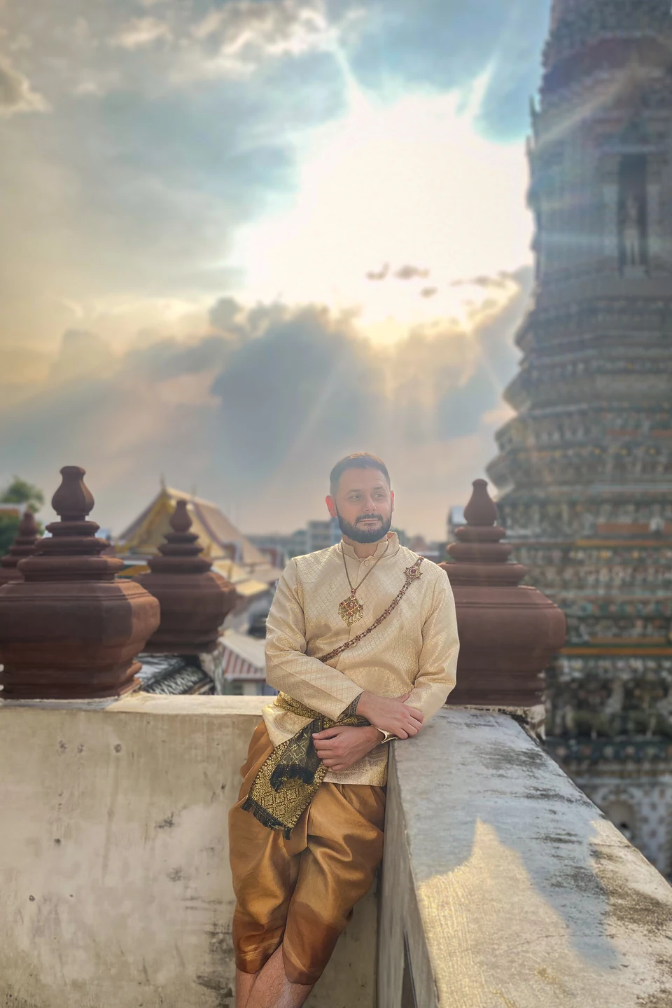 Peter Zošiak stojí opřený o zeď v tradičním thajském oblečení se zlatým zdobením. V pozadí je historický chrám Wat Arun v Bangkoku, slunce prosvítá mezi mraky a vytváří dramatickou atmosféru.