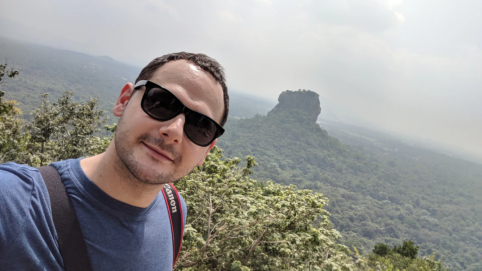 Peter Zošiak pořizuje selfie s ikonickou skalní pevností Sigiriya na Srí Lance v pozadí. Má na sobě tmavé sluneční brýle, modré tričko a popruh od fotoaparátu přes rameno.