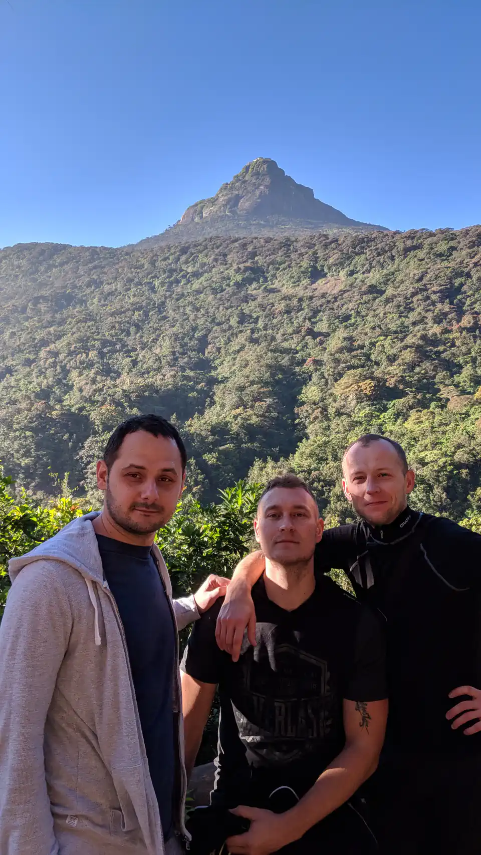 Peter Zošiak se dvěma přáteli stojí před horou Adam’s Peak na Srí Lance. Všichni jsou oblečeni do sportovního oblečení, slunce osvětluje zelené svahy hory v pozadí.