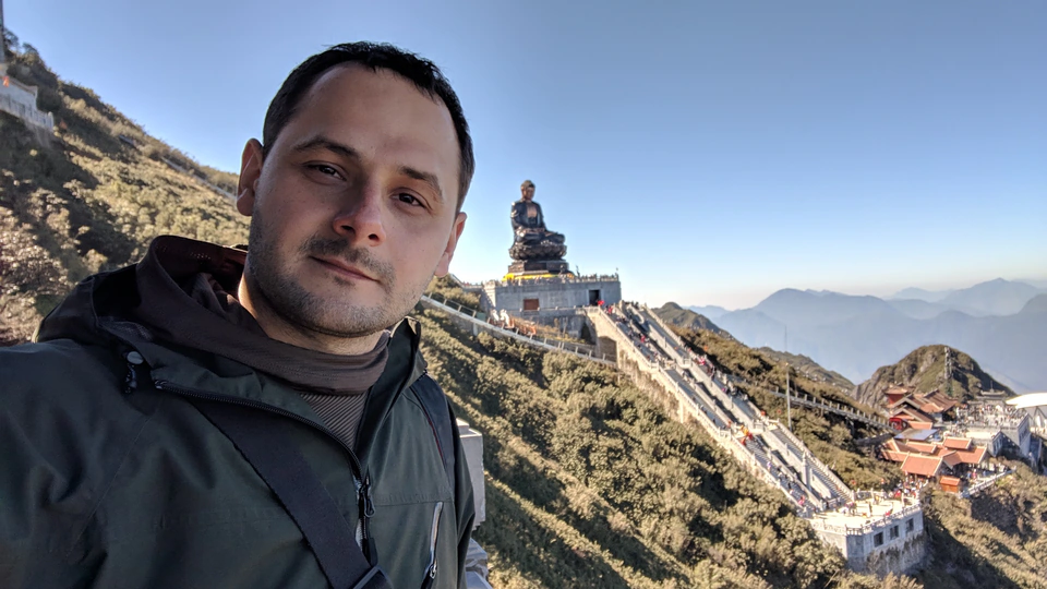 Peter Zošiak pořizuje selfie na horském hřebeni s obřím Buddhou a schodištěm vedoucím k chrámu v pozadí ve Vietnamu na Fansipan v Sapa. Jasná obloha a horské panorama dodávají scéně majestátní atmosféru.