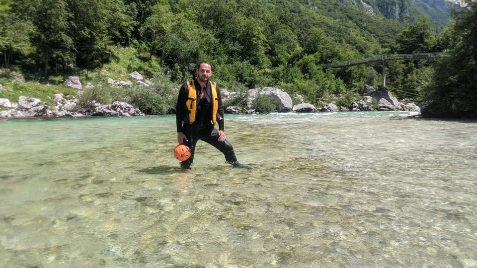 Peter Zošiak stojí v mělké řece po adrenalinovém canyoningu v Slovinsku na řece Soča. Má na sobě černou neoprenovou kombinézu, žlutou vestu a drží helmu. Okolní scenérie je plná zelených lesů a kamenů.
