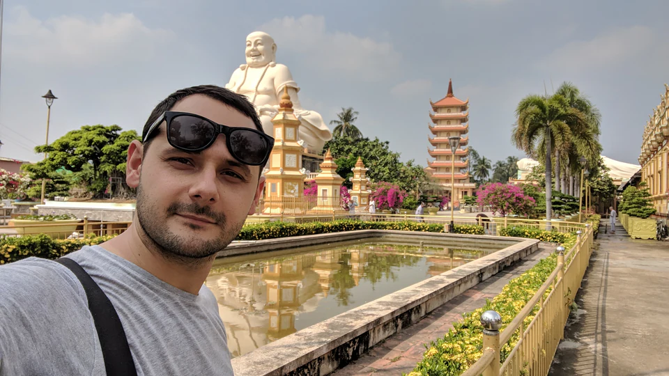 Peter Zošiak se fotí před zlatou sochou smějícího se Buddhy a pagodami ve vietnamském chrámovém komplexu. Okolí zdobí jezírko, palmy a květiny.
