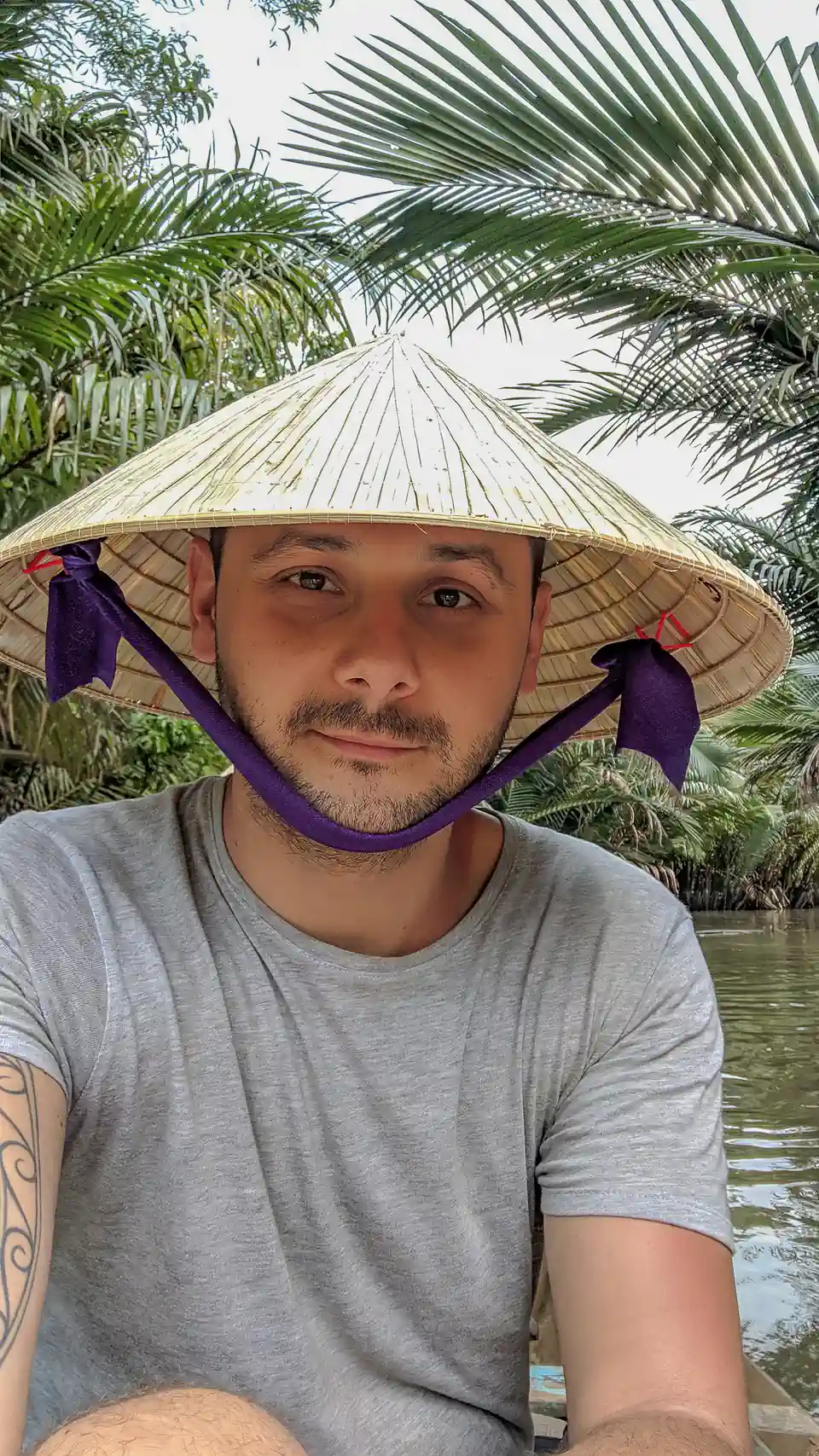 Peter Zošiak v tradičním vietnamském klobouku „nón lá“ sedí na lodi uprostřed tropických palem při plavbě po řece Mekong.