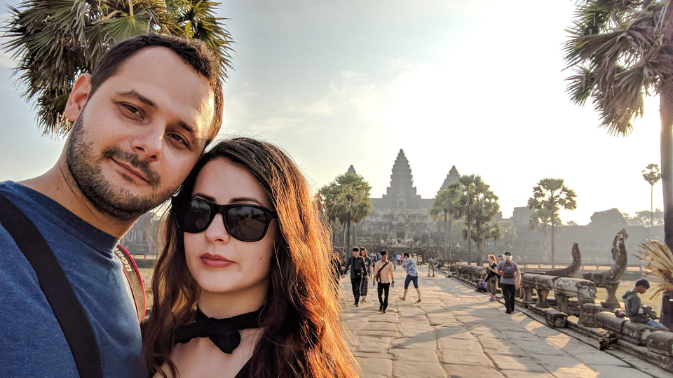 Peter Zošiak a jeho partnerka pózují před ikonickým chrámem Angkor Wat v Kambodži při západu slunce. Kolem nich jsou palmy a turisté obdivující historickou památku.